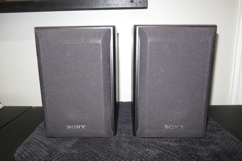 May include: A pair of rectangular black Sony speakers featuring dark grey fabric grilles covering the front panels. Each speaker has the Sony brand name printed in silver lettering at the bottom center of the front face, with a solid black frame.