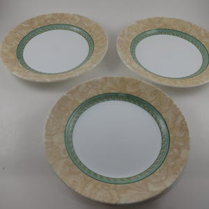 May include: Three white ceramic plates with a beige and green decorative border. The plates have a white center and a beige border with a subtle, swirling pattern. The green border is a thin band that adds a pop of color.