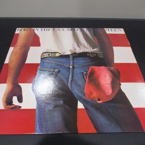 May include: A vinyl record album cover featuring a man wearing blue jeans and a white t-shirt, standing in front of an American flag. The album title "Born in the U.S.A." and the artist's name "Bruce Springsteen" are printed on the cover.