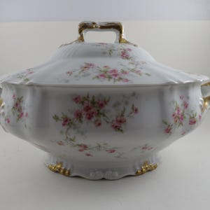 May include: White porcelain serving dish with lid, adorned with pink floral patterns. Features gold trim on the handles, base, and lid handle. A classic piece for serving soups or stews.