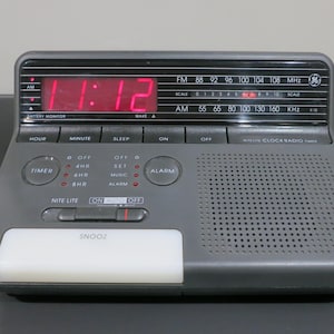 May include: A gray digital clock radio with a red LED display showing 11:12. It has buttons for hour, minute, sleep, and alarm, with a snooze bar and a radio frequency scale. The clock radio is a vintage model.