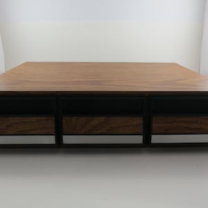 May include: A wooden audio component cabinet with three compartments. The top is a light brown wood grain, and the front has black vents and silver trim. The cabinet is rectangular and sits on a white surface.