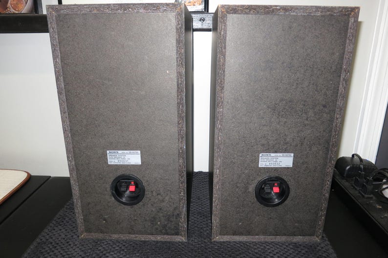 May include: Two rectangular Sony speakers with dark grey textured surfaces. Each speaker has a black circular port with red terminals and a white label. The speakers are positioned side by side.