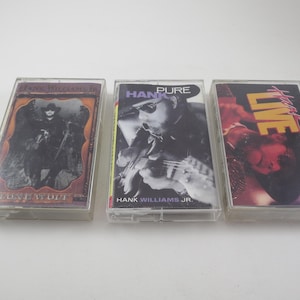 May include: Three cassette tapes with different album covers. The first tape has a black and white image of a man in a cowboy hat and a long coat. The second tape has a color image of a man with a guitar. The third tape has a color image of a man with a microphone. The text on the tapes reads "The Outlaw in Me", "Hank Pure", and "Live".