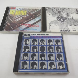 May include: Three compact discs featuring album art from The Beatles. The top left CD is "Please Please Me", the top right is "Revolver", and the bottom CD is "A Hard Day's Night". All three CDs are in clear plastic cases.