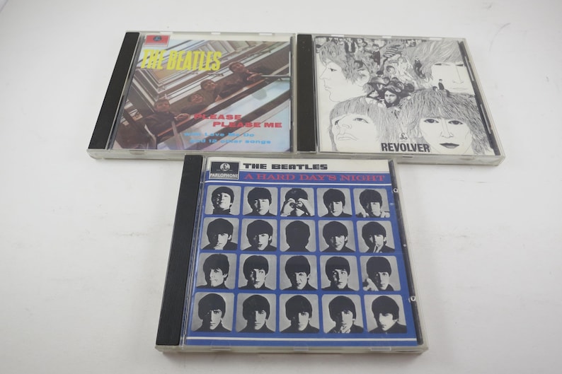 May include: Three compact discs featuring album art from The Beatles. The top left CD is "Please Please Me", the top right is "Revolver", and the bottom CD is "A Hard Day's Night". All three CDs are in clear plastic cases.