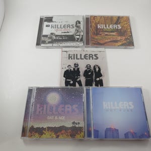 May include: A collection of five CDs by the band The Killers. The album covers feature various designs, including band photos, landscapes, and text. The CDs are in clear plastic cases, with the band's name prominently displayed.