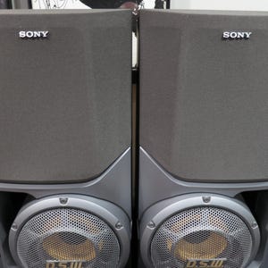 May include: Two gray Sony speakers. Each speaker has a rectangular, fabric-covered upper section with the "SONY" logo. The lower section features a silver mesh grill over a circular speaker, with "D.S.W." printed on it.