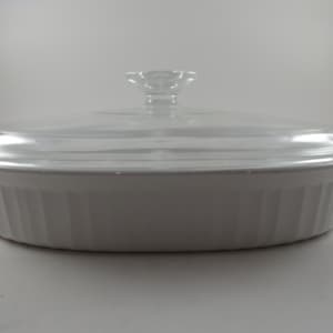 May include: Oval white casserole dish with a clear glass lid. The dish has a ribbed design and a clear glass lid with a flower-shaped handle. A kitchenware item for baking and serving.