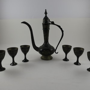 May include: An antique dark metal tea set with a tall, ornate pitcher and six matching goblets. The pitcher has a long spout and a handle. The goblets are small and have a decorative pattern. The set is likely for serving tea or other beverages.