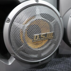 May include: Close-up of two silver speakers with a perforated metal grill. The center of the speaker is gold. The text "D.S.III" is visible on the speaker. The speakers are part of a larger audio system.