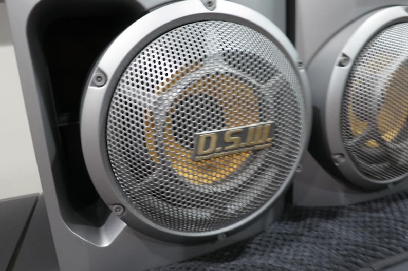 May include: Close-up of two silver speakers with a perforated metal grill. The center of the speaker is gold. The text "D.S.III" is visible on the speaker. The speakers are part of a larger audio system.