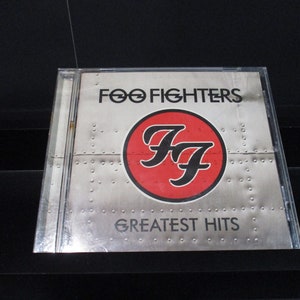 May include: A Foo Fighters "Greatest Hits" CD in a clear plastic case. The album cover features the band's logo, a red circle with the letters "FF" in black, and the band's name above. The background is a silver, riveted metal design.