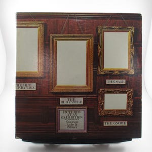 May include: A vintage record album cover featuring a wooden wall with five framed pictures. The album is titled "Pictures at an Exhibition" by Emerson, Lake & Palmer. The album cover features the text "The Old Castle", "The Hut Baba Yaga", "The Sage", and "The Gnome".
