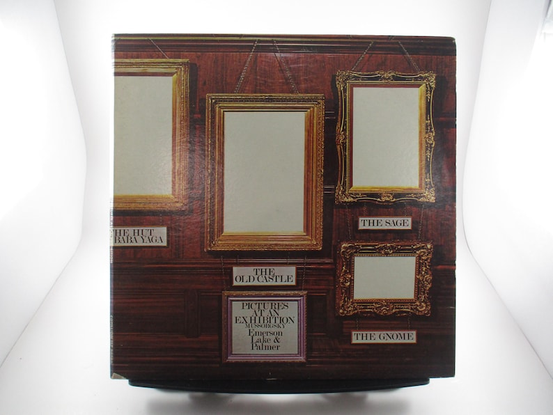 May include: A vintage record album cover featuring a wooden wall with five framed pictures. The album is titled "Pictures at an Exhibition" by Emerson, Lake & Palmer. The album cover features the text "The Old Castle", "The Hut Baba Yaga", "The Sage", and "The Gnome".