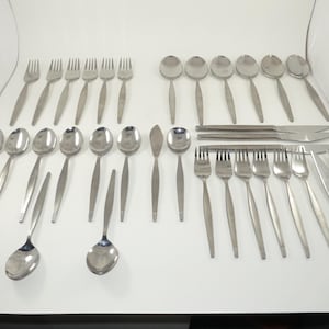 May include: A large collection of stainless steel flatware including various forks, spoons, and knives. The set features a simple, smooth design with tapered handles. The pieces are arranged in rows on a white surface, showcasing a complete set of dining utensils for multiple place settings.
