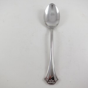 May include: A silver spoon with a rounded bowl and a long, slender handle. The handle flares out slightly near the base, with an ornate, decorative design. The spoon is made of a shiny, reflective metal.