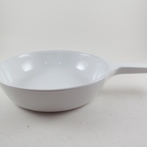 May include: A white ceramic skillet with a single handle. The skillet has a rounded bowl shape and a smooth, glossy finish. The handle is rectangular and extends horizontally from the side of the bowl. The skillet is designed for cooking and serving.