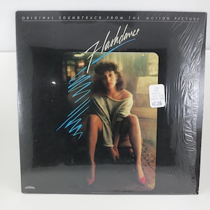 May include: Sealed vinyl record of the "Flashdance" soundtrack. The album cover features a woman in a seated pose, with the title in a stylized font and blue accents. The album is in its original packaging.