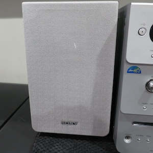 May include: A silver and gray Sony stereo system with a speaker and a control panel. The speaker is rectangular with a gray fabric cover and the Sony logo. The control panel has buttons and a digital display.