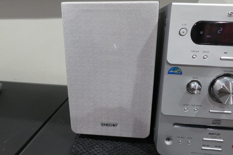 May include: A silver and gray Sony stereo system with a speaker and a control panel. The speaker is rectangular with a gray fabric cover and the Sony logo. The control panel has buttons and a digital display.