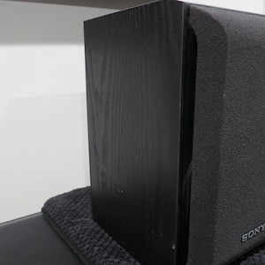 May include: A pair of black Sony bookshelf speakers featuring a textured wood grain finish on the side panels and a dark fabric grille covering the front. The Sony brand name is printed in small silver letters at the bottom of the front grille.
