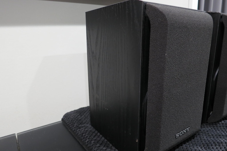 May include: A pair of black Sony bookshelf speakers featuring a textured wood grain finish on the side panels and a dark fabric grille covering the front. The Sony brand name is printed in small silver letters at the bottom of the front grille.