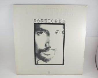 May include: A vintage vinyl record album cover featuring a black and white portrait of a man's face. The album title "FOREIGNER" is printed in bold, black letters, with the artist's name embossed above. The cover is a light cream color.