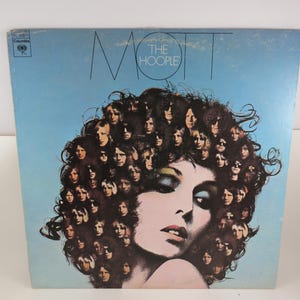May include: A square vinyl record cover with a light blue background. The text MOTT THE HOOPLE is printed at the top. The center features a large illustration of a face surrounded by a collage of many smaller faces forming a hair-like shape.
