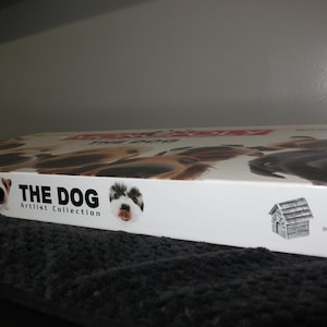 May include: A white Monopoly The Dog Artist Collection board game box featuring various dog portraits on the lid. The side panel displays the text THE DOG Artist Collection, images of two dogs, a graphic of a dog house, and 6 Custom Pewter Tokens.