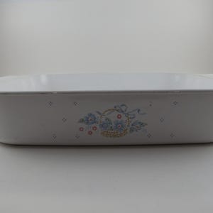 May include: Rectangular white ceramic baking dish with a floral basket design. The dish has a light blue and red floral pattern with a basket of flowers and blue ribbons. The dish has a silver trim and is approximately 13 inches long.
