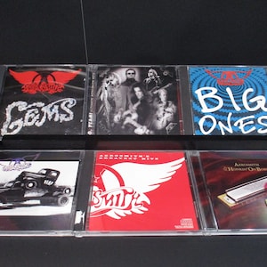 May include: Six compact discs in clear plastic cases, featuring various rock bands. The discs include Aerosmith's "Gems", "Big Ones", "Greatest Hits", "Honkin' On Bobo", and two other Aerosmith albums.