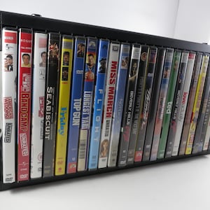 May include: Black DVD storage rack filled with various movie titles. Titles include "Band Camp," "Seabiscuit," "Top Gun," and "Miss March." The rack is angled, showcasing the DVD cases. The rack is approximately 12 inches wide.