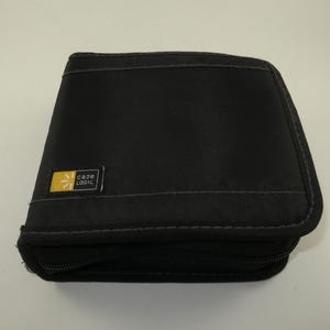 May include: A black zippered media storage case with a square shape and rounded corners. The exterior is made of a durable fabric material. A small rectangular tag with a yellow sun icon and the text Case Logic is attached to the front cover.