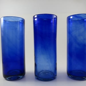 May include: Three tall, cylindrical drinking glasses in a vibrant cobalt blue hue. The glasses have a slightly rounded top and a smooth, transparent texture, with subtle variations in color. They are evenly spaced against a plain white background.