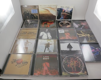 May include: A collection of Neil Young CDs, featuring album art with various designs and colors. The CDs are in clear jewel cases, showcasing the album covers. Some covers feature photographs, while others have illustrations and text.