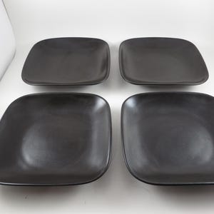 May include: Four square, dark gray plates with rounded corners are arranged on a white surface. The plates have a matte finish and a slightly concave design. The plates are a uniform color and shape.