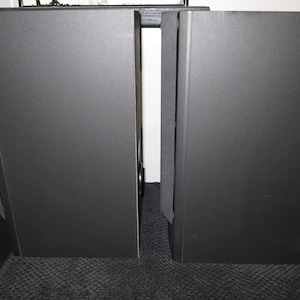 May include: Two black rectangular speakers with a textured surface stand side by side. The speakers are tall and narrow, with a simple, modern design. The image is well-lit, highlighting the speakers' dark colour and shape. The speakers are likely for audio equipment.