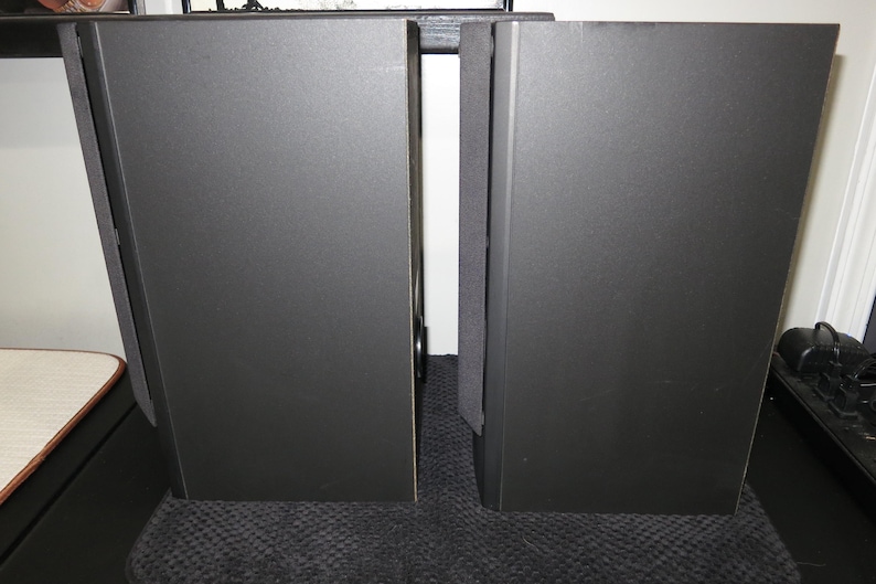 May include: Two black rectangular speakers with a textured surface stand side by side. The speakers are tall and narrow, with a simple, modern design. The image is well-lit, highlighting the speakers' dark colour and shape. The speakers are likely for audio equipment.