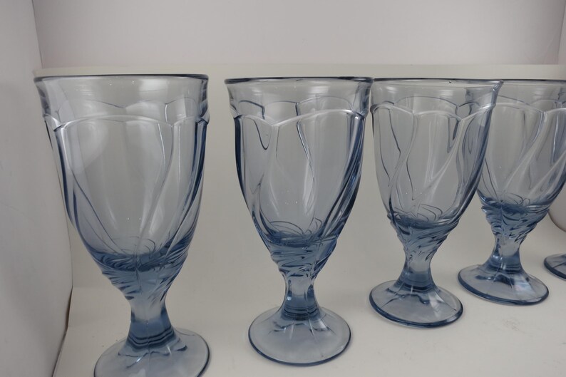 Pu&ograve; includere: Un set di calici in vetro azzurrato. Ogni calice presenta una coppa svasata con un motivo a spirale decorativo e un gambo. I bicchieri sono disposti in fila, mostrando la loro forma e il loro colore eleganti.