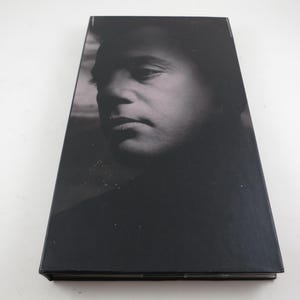 May include: A black and white image of a book cover featuring a portrait of a man. The man's face is in the center of the cover, with his eyes looking down. The cover is black and glossy.