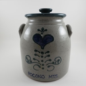 May include: A gray ceramic jar with a lid and two handles. The jar is decorated with a blue heart and floral design. The text "POCONO MTS." is written on the bottom of the jar.