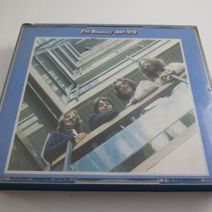 May include: A CD case featuring the album cover of The Beatles' 1967-1970 compilation. The cover art shows the band members leaning over a balcony. The case has a blue border and the album title is printed at the top.