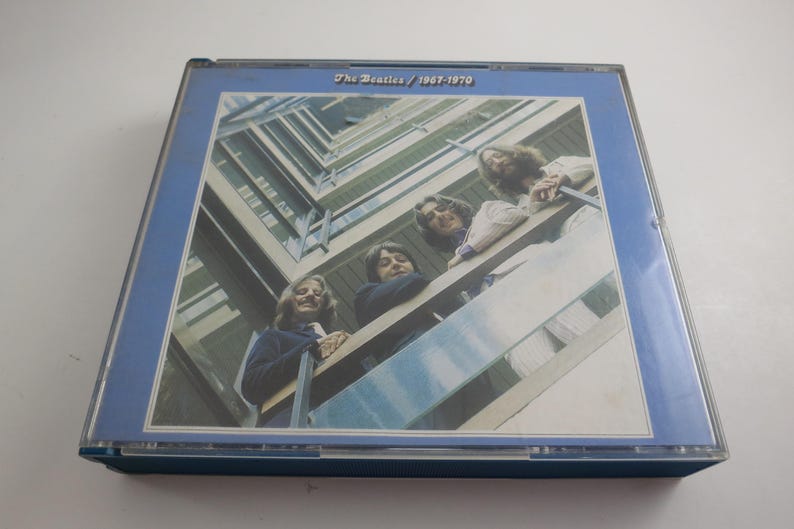 May include: A CD case featuring the album cover of The Beatles' 1967-1970 compilation. The cover art shows the band members leaning over a balcony. The case has a blue border and the album title is printed at the top.