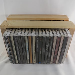 May include: A light-colored wooden crate containing a row of twenty-one compact disc cases arranged vertically. The spines of the cases display various artist names and album titles in black, white, and colored text against dark backgrounds.