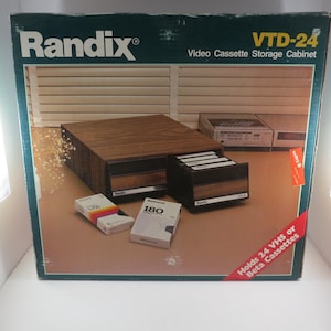 May include: A Randix VTD-24 video cassette storage cabinet. The cabinet is made of wood and has two drawers. The cabinet holds 24 VHS or Beta cassettes. The box also contains three video cassettes.