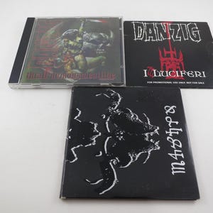 May include: Three music albums are displayed. One is a CD in a clear case with a demonic illustration. The other two are black with white text and illustrations. One reads "DANZIG I Luciferi". The other reads "m437p8".