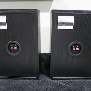 May include: Two black Sony speaker cabinets shown from the rear, each featuring a white product information label at the top and a circular connection terminal with one red and one black spring-loaded clip in the center of the wooden housing.
