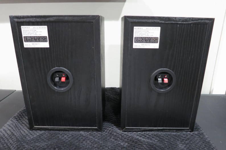 May include: Two black Sony speaker cabinets shown from the rear, each featuring a white product information label at the top and a circular connection terminal with one red and one black spring-loaded clip in the center of the wooden housing.