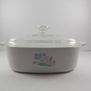 May include: A rectangular, off-white casserole dish with a clear glass lid. The dish features a floral design in blue, pink, and yellow. The lid has a clear, flower-shaped handle. This vintage dish is perfect for baking and serving.